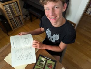 a young man working at a table with a pile of books, smiling up at the camera