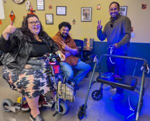 A smiling woman with a halloween party hat, sitting on a travelscoot, with two guys doing thumbs up behind her