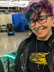 white non binary person with glasses and purple hair, smiling, wearing tshirt with wheelchair user with symbolic flames trailing behind them