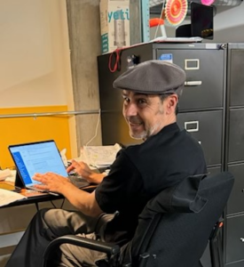 A bearded man smiling at a desk, wearing a flat cap